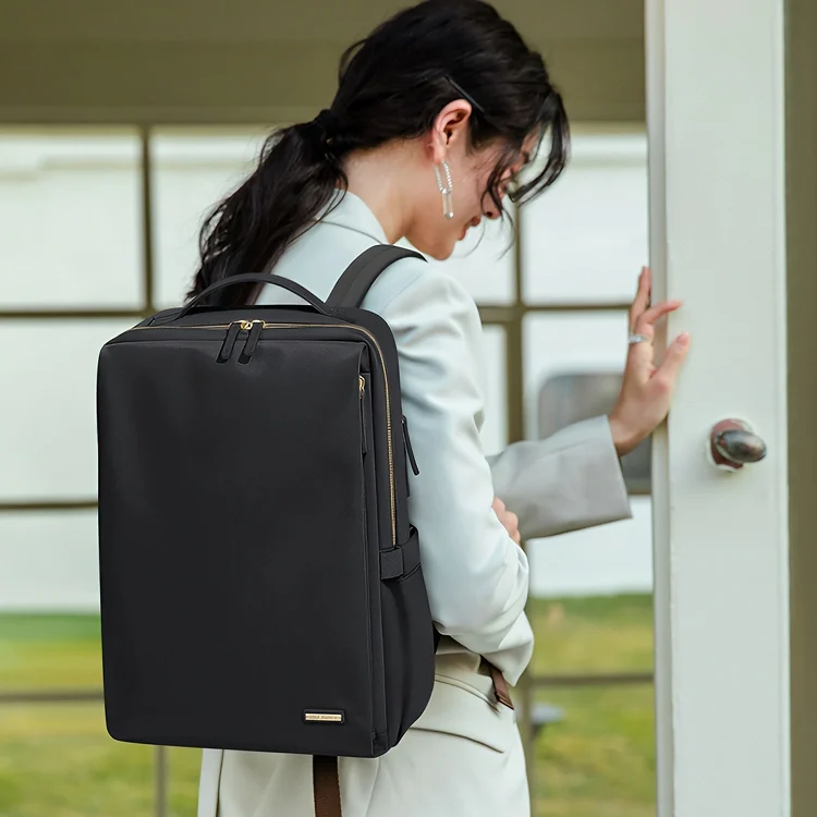 黒のビジネスリュックを背負い、白いジャケットを着た女性がドアに手をかけている姿。リュックは15.6インチ対応で多収納機能が特徴。