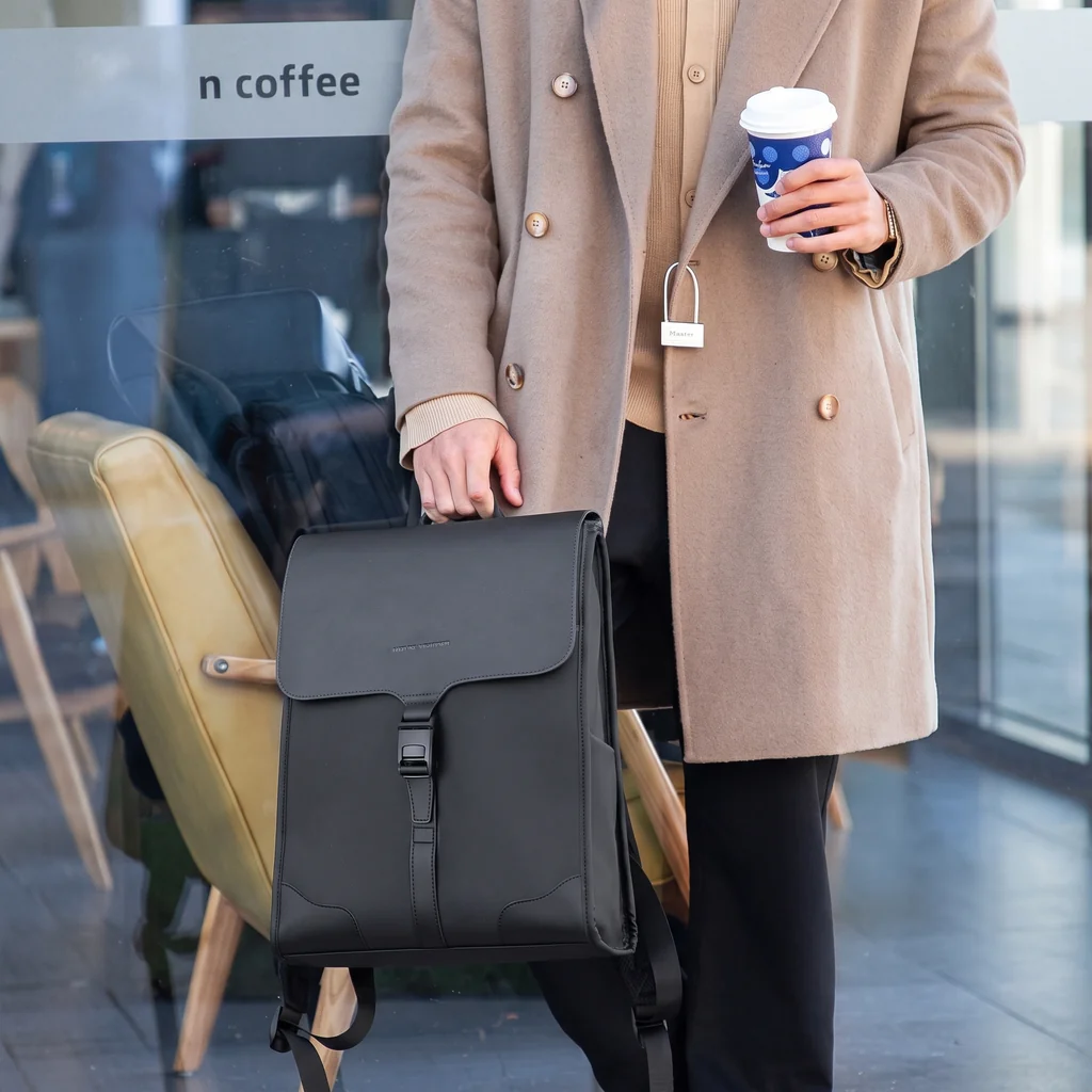 シックなトレンチコートを着た男性が持つ、自立式で撥水性のあるおしゃれな20Lスクエアバックパック。カフェの前で佇む洗練された様子。