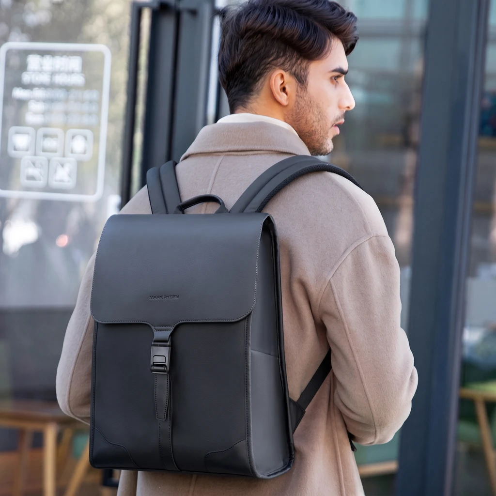 ベージュのオーバーコートを着た男性が、撥水仕様の20L自立型スクエアバックパックを背負って都会のモダンなカフェの前に立つ様子