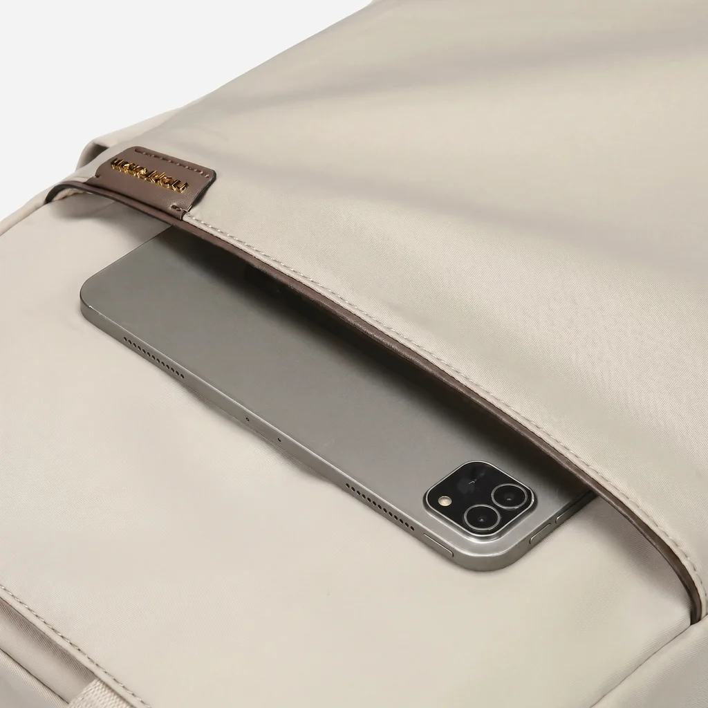 上品なベージュのレディース防水ビジネスリュックのフロントポケットへ、グレーのタブレットをスマートに収納する手元のクローズアップ。