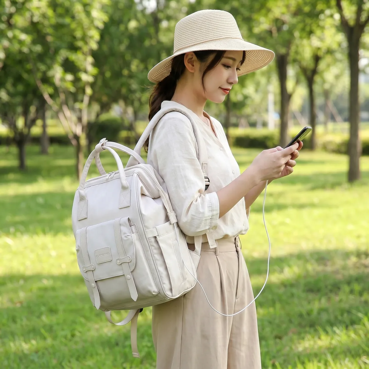 陽光が差し込む緑豊かな公園で、kireimeの多機能防水マザーズリュックに接続したスマホを操作する、麦わら帽子とリネン服の女性。