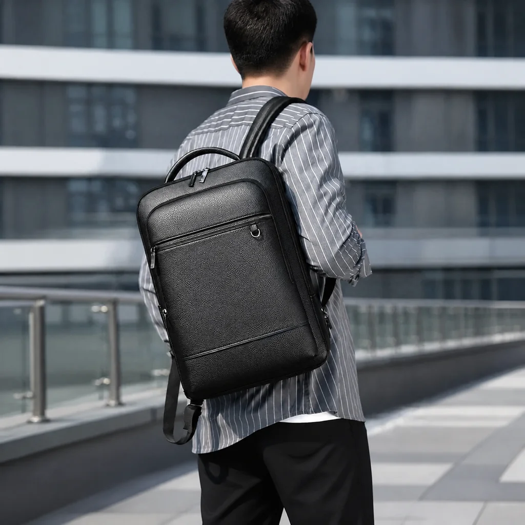 グレーのストライプシャツを着て、16インチPCが入る20L多機能ビジネスリュックを背負い、近代的なオフィス街を歩く男性の後ろ姿。