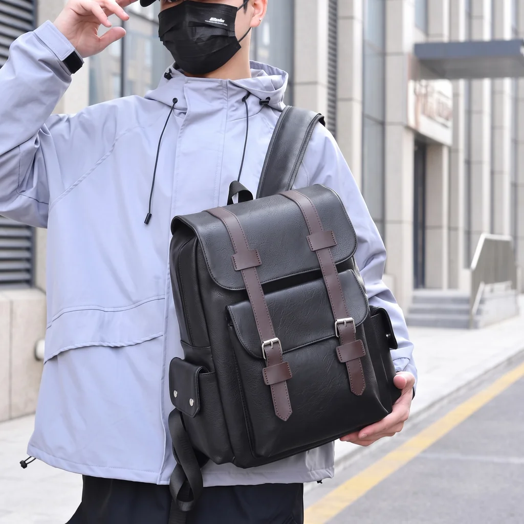 グレーのジャケットを着た男性が手にする、20L防水仕様のメンズ用レトロレザーバックパック。洗練されたモダンな都会の街並み。