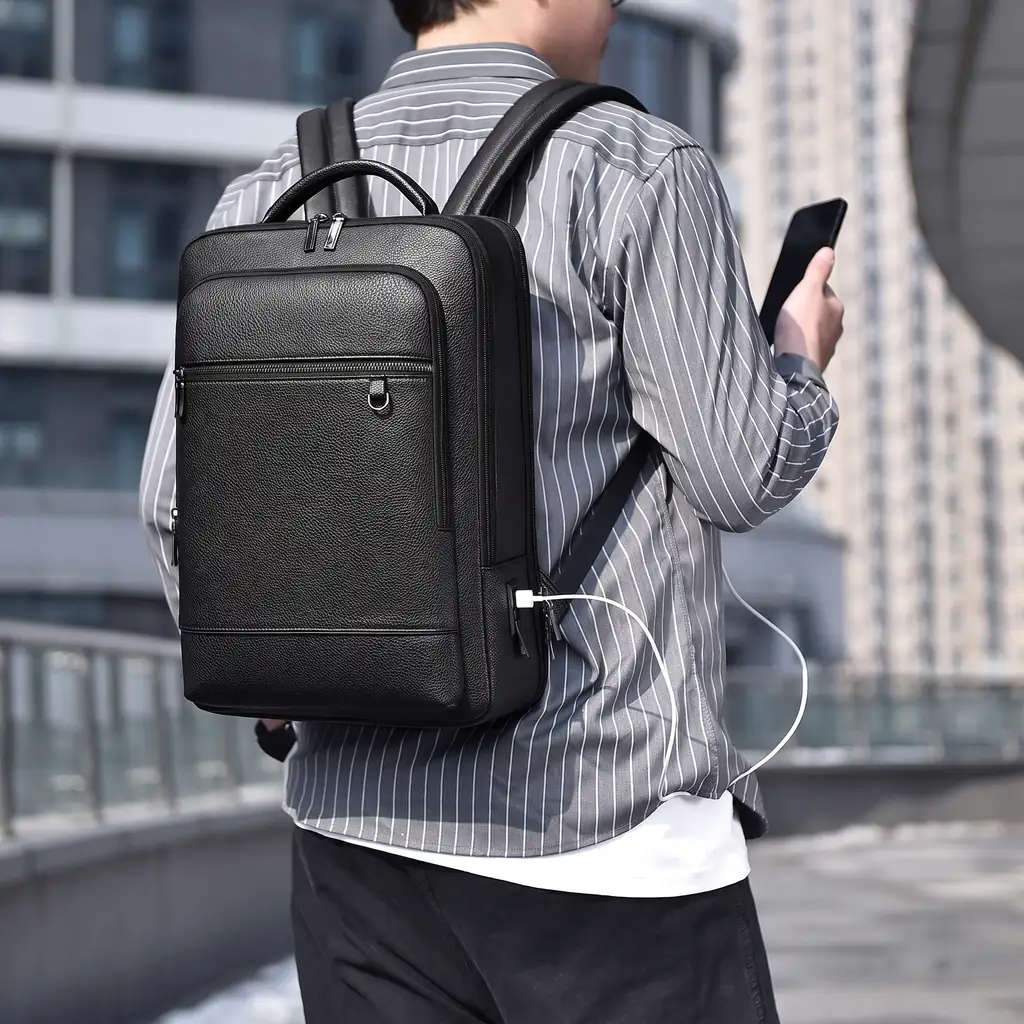 高層ビル街を歩くグレーのシャツを着た男性が背負う、充電ケーブルを繋いだスマホを持つ、16インチPC対応20L多機能ビジネスリュック。