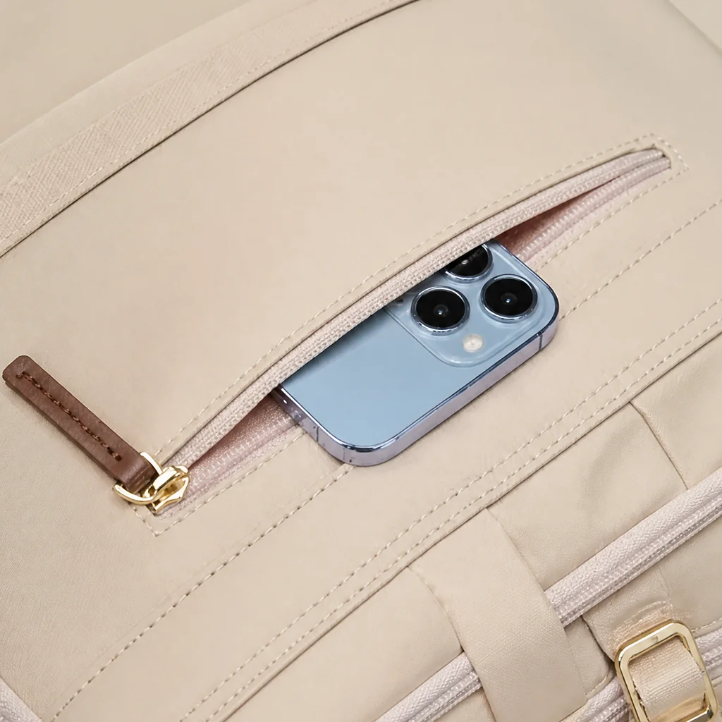 レディースリュックの背面ファスナーポケットとスマホ収納 ベージュのレディースリュックの背面ファスナーポケットに青いスマホを収納する様子。ブラウンの革製引き手がおしゃれな背面収納のクローズアップ。