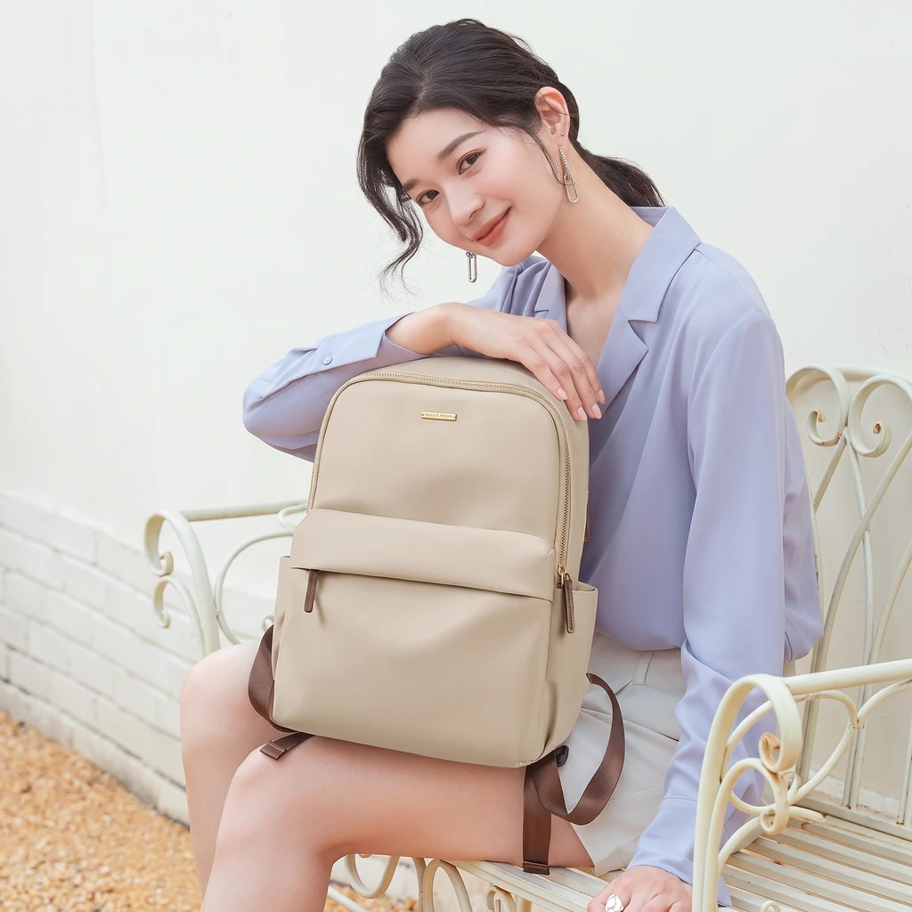 明るい光が差し込むテラスの白いベンチに座り、A4対応の軽量撥水ベージュレディースリュックを膝に置くおしゃれな女性の姿。