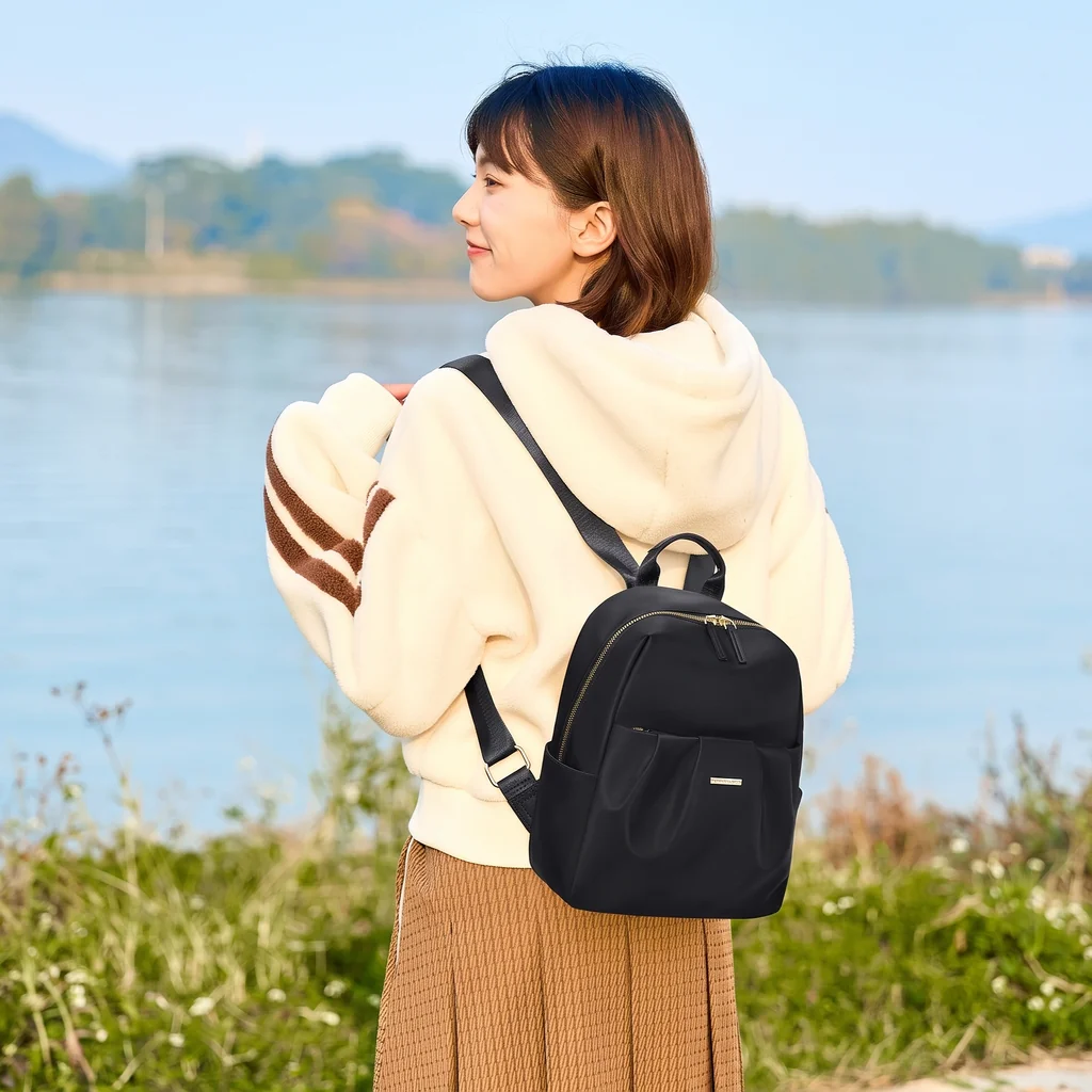 穏やかな湖畔の芝生に立ち、女性用の小さなアーバンスタイリッシュバックパックを背負って振り返る、クリーム色のパーカーを着た女性。