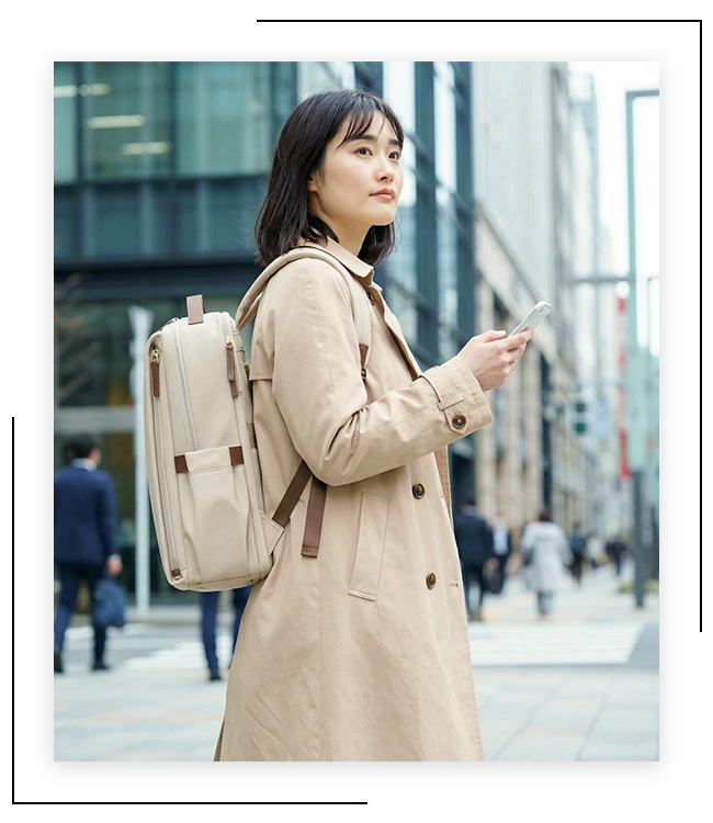 ビル街でスマートフォンを手に歩く、ベージュのトレンチコートに都市型デイリーリュックを背負った女性