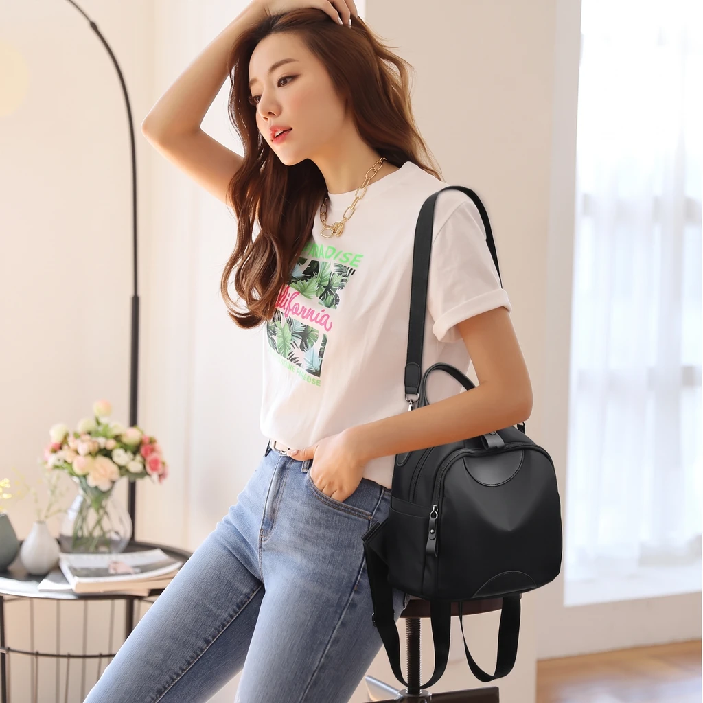 明るい部屋で撥水ナイロン製レディース15Lミニリュックを肩に掛けるカジュアルな女性。窓からの自然光とピンクの花が飾られたおしゃれな空間。